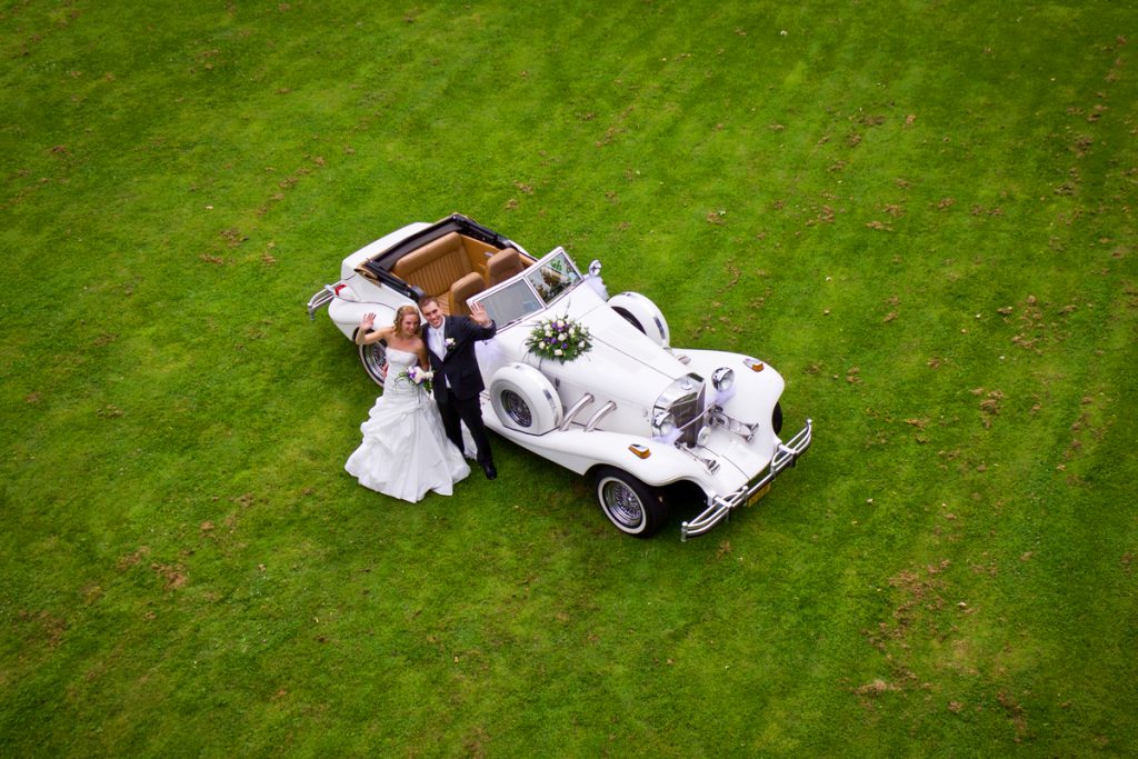 Luchtfoto tijdens bruidsreportage van koppel bij de auto op een grasveld