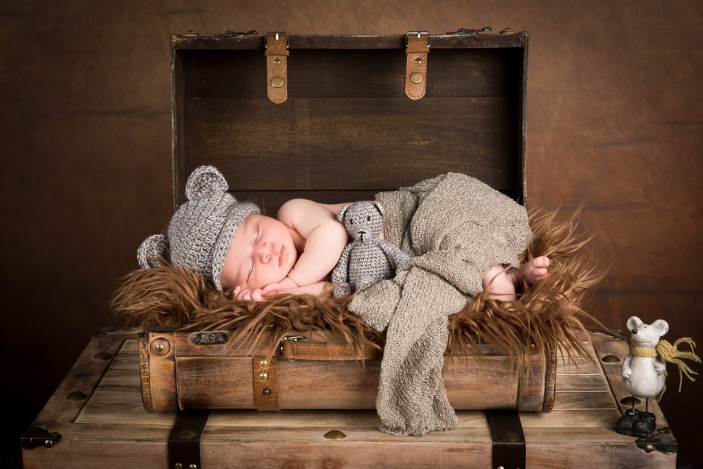 Baby Djévano slapend in een kist gewikkeld in een bruine deken met een grijs mutsje op en een grijs beertje erbij