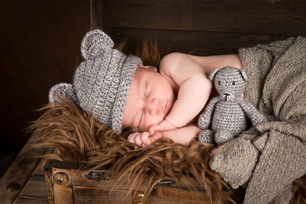 Baby Djévano slapend in close-up in een kist gewikkeld in een bruine deken met een grijs mutsje op en een grijs beertje erbij