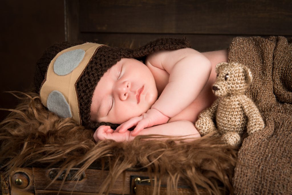 Baby Djévano slapend in close-up slapen in een kist met een vliegeniersmuts op en gewikkeld in een bruine deken met een bruin beertje erbij