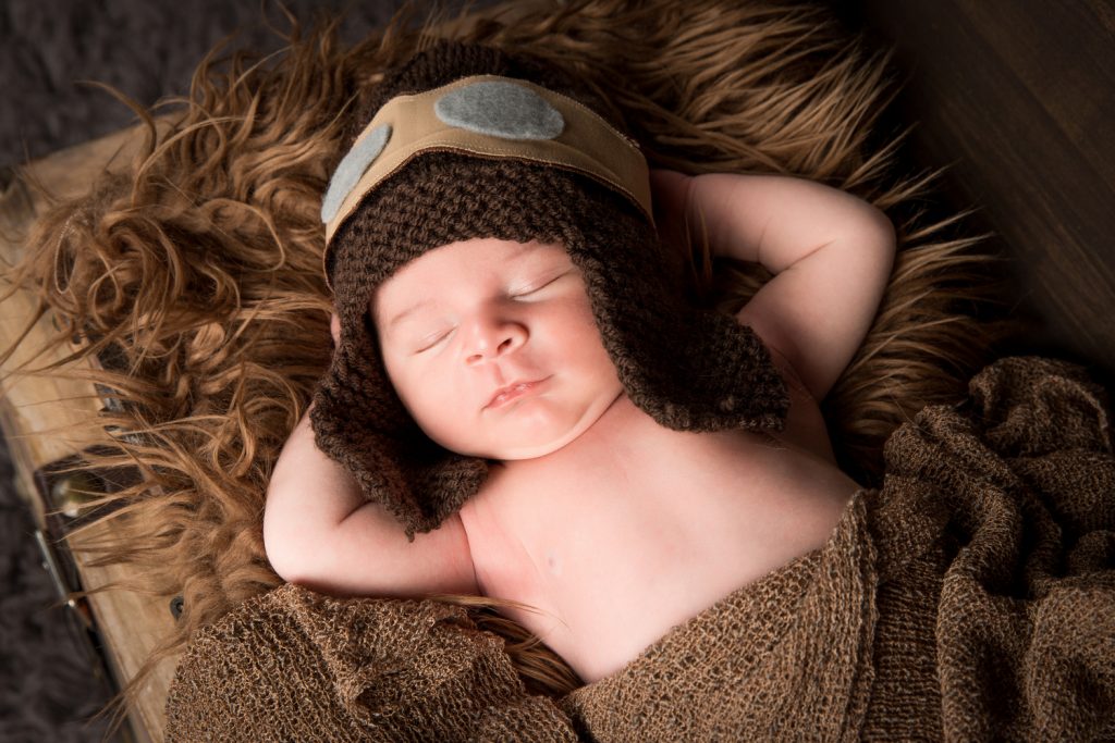 Baby Djévano slapend in close-up slapend in een kist met zijn armen onder zijn hoofd en een vliegeniersmuts op en gewikkeld in een bruine deken