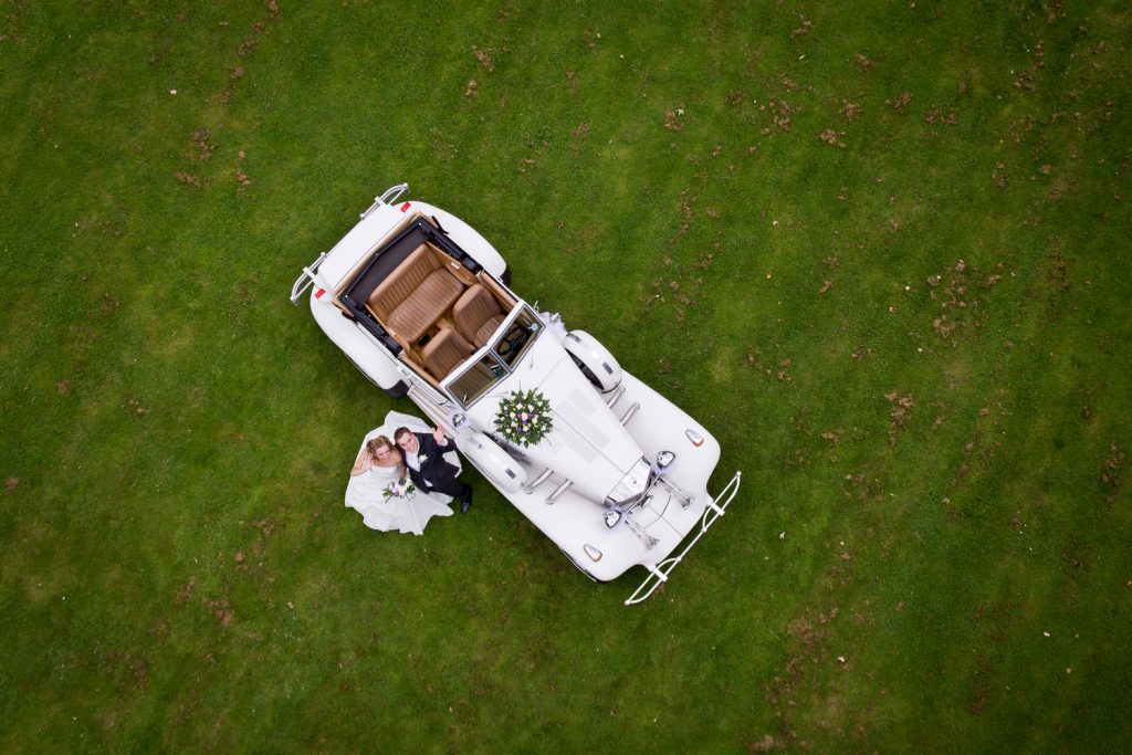Luchtfoto tijdens bruidsreportage van koppel bij de auto op een grasveld